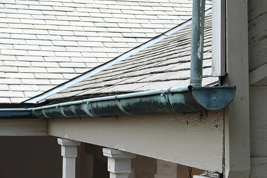 Copper-colored gutter and downspout below a slate roof edge and white trim